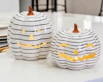 May include: Two white and black striped ceramic pumpkin candle holders with a jack-o-lantern face design. The pumpkins are lit from inside with a warm glow.