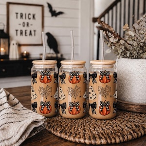 May include: Three glass tumblers with wooden lids and clear straws. Each tumbler is decorated with a pumpkin, spiderweb, ghost, and bow design. The background includes a sign that says "Trick or Treat".