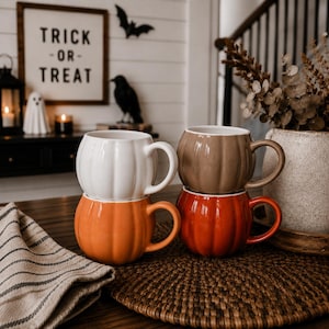 Może przedstawiać: Cztery ceramiczne kubki w kształcie dyń, ułożone w dwie pary. Kubki mają żebrowany, zaokrąglony kształt z uchwytem z boku. Kolory obejmują biały, beżowy, pomarańczowy oraz ciemny czerwono-pomarańczowy, wszystkie z błyszczącym wykończeniem.