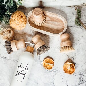 May include: A white bottle of dish soap with black lettering that reads "dish soap" and "CIA ROMA". The bottle is surrounded by wooden dish brushes and sponges on a white countertop.