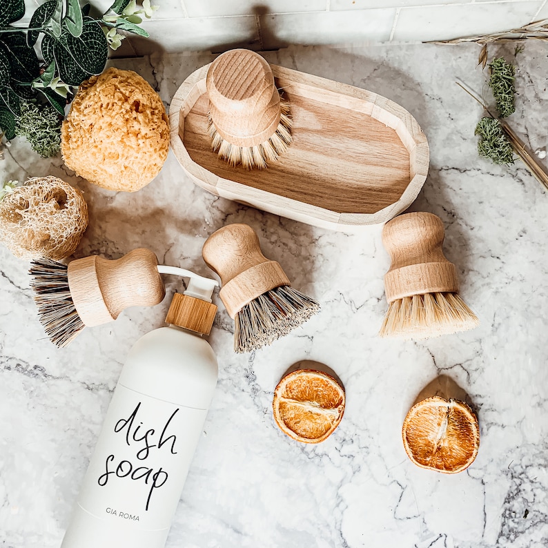 May include: A white bottle of dish soap with black lettering that reads "dish soap" and "CIA ROMA". The bottle is surrounded by wooden dish brushes and sponges on a white countertop.