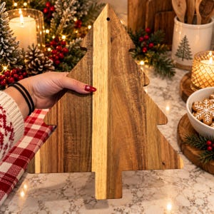 May include: A Christmas tree-shaped wooden serving board featuring a vertical light-colored wood grain stripe down the center, flanked by darker brown wood grain panels on both sides, with a small circular hole at the top point for hanging.