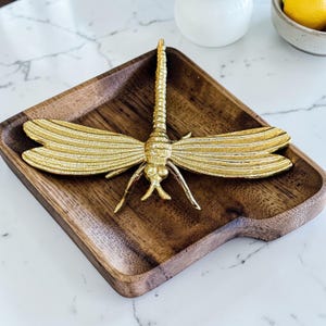 May include: A decorative gold-colored dragonfly figurine rests on a square wooden tray. The dragonfly has detailed wings and body. The tray is made of dark wood and has rounded edges. The background is a white marble surface.