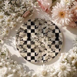 Op de afbeelding: Een ronde decoratieve plaat met een zwart-wit geblokt patroon, bedekt met een bloemenontwerp in wit- en cr&egrave;metinten. De plaat is omgeven door witte en roze bloemen.