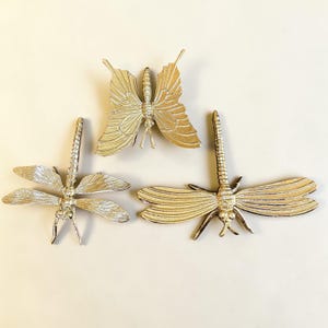 May include: Three decorative metal insect sculptures: a butterfly and two dragonflies. The butterfly has textured wings and a detailed body. The dragonflies have detailed wings and bodies. All are gold-colored.