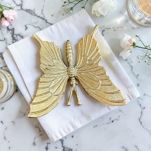 May include: A gold-colored butterfly-shaped decorative object with detailed wing patterns, resting on a white folded cloth. The butterfly is centered in the frame, with a marble background and floral accents.