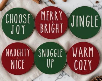 May include: A set of six round drink coasters in red and dark green with white text. The coasters feature the phrases CHOOSE JOY, MERRY + BRIGHT, JINGLE, NAUGHTY + NICE, SNUGGLE UP, and WARM + COZY in a simple, hand-lettered style font.