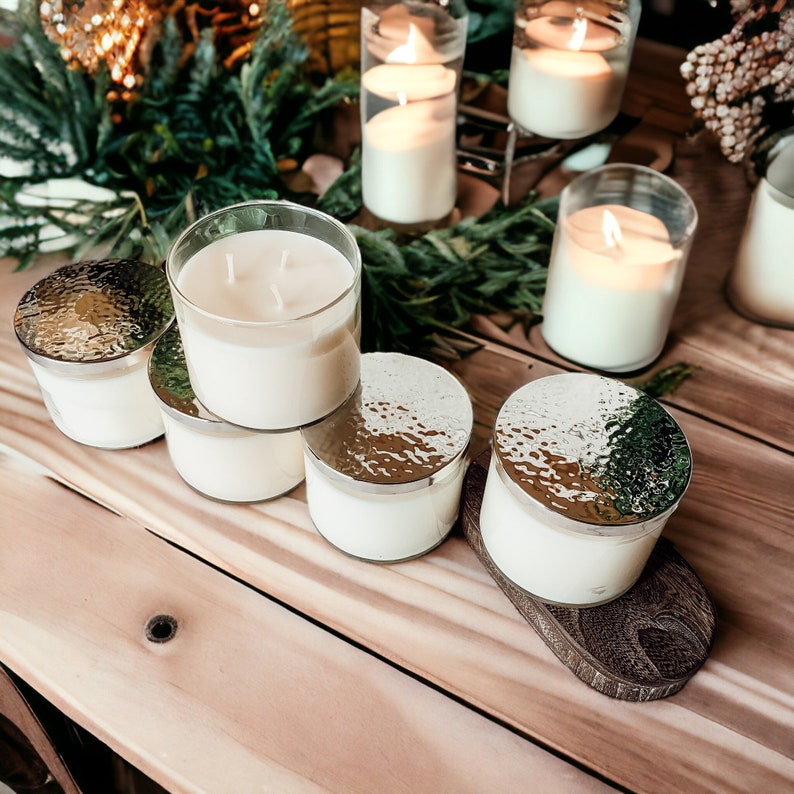 May include: A collection of six candles in glass jars with silver lids. The candles are arranged on a wooden surface and are lit. The candles are surrounded by greenery.