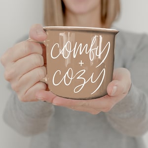 Puede incluir: Una taza de esmalte marrón con las palabras "comfy + cozy" en escritura blanca. La taza tiene un borde blanco y un asa. La taza es sostenida por una persona que lleva una camiseta gris de manga larga.