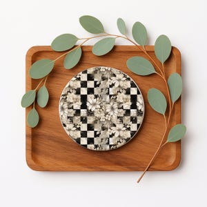 Op de afbeelding: Een ronde onderzetter met een zwart-wit geblokt patroon, bedekt met een bloemenontwerp in tinten wit en grijs. De onderzetter ligt op een houten dienblad, met groene eucalyptusbladeren.