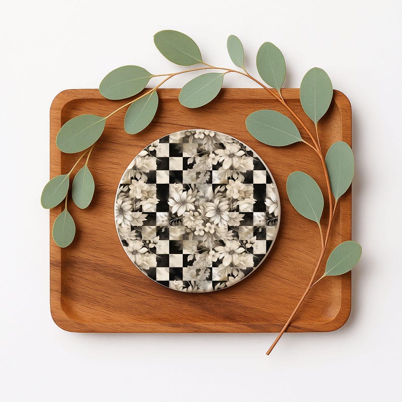 Op de afbeelding: Een ronde onderzetter met een zwart-wit geblokt patroon, bedekt met een bloemenontwerp in tinten wit en grijs. De onderzetter ligt op een houten dienblad, met groene eucalyptusbladeren.