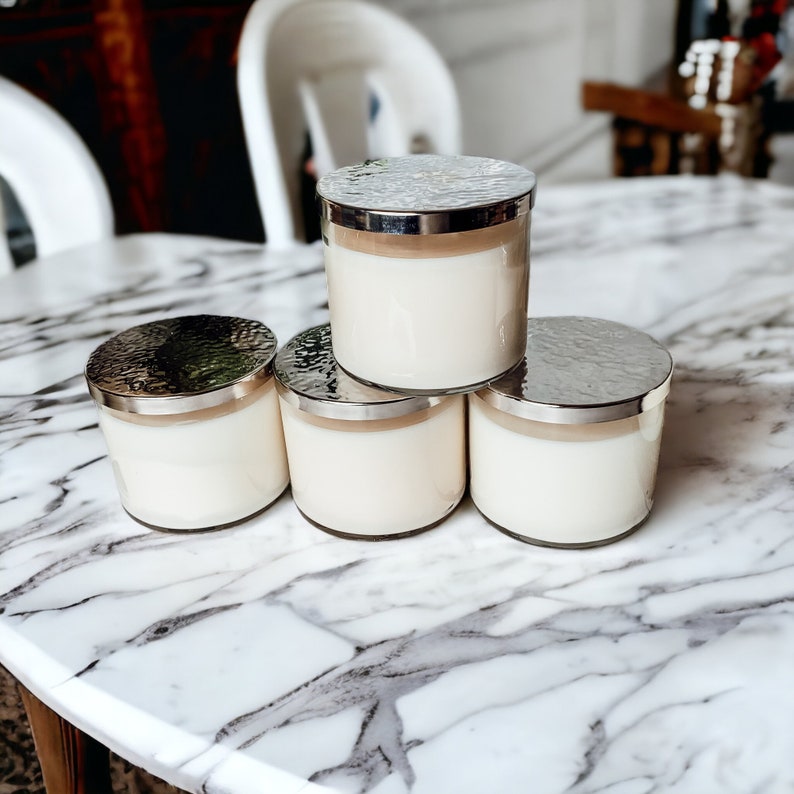 May include: Four white candles with silver lids are stacked on a white marble table. The candles are in glass jars with a hammered silver finish.