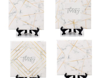 May include: Four white ceramic coasters with gold geometric designs and marble patterns. Each coaster has a black metal stand and the word "thirsty" written on two of the coasters.