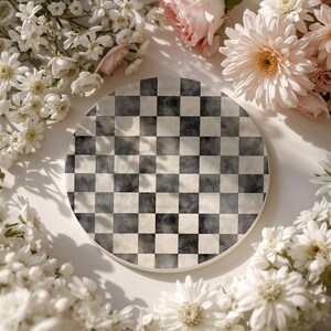 Op de afbeelding: Een ronde onderzetter met een zwart-wit dambordpatroon. De onderzetter is omgeven door witte en roze bloemen, wat een zachte, elegante uitstraling cre&euml;ert. Het ontwerp doet denken aan een schaakbord.