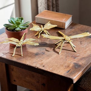 May include: Three gold-colored dragonfly sculptures with outstretched wings are displayed on a wooden table. A small potted succulent and a closed book are also on the table. The dragonflies have detailed wings and slender legs, adding a touch of nature-inspired decor.