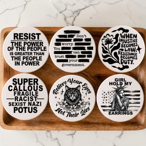 Può includere: Sei sottobicchieri in ceramica bianca con testo e disegni neri. I sottobicchieri presentano varie dichiarazioni e grafiche politiche, tra cui il testo che recita "RESIST THE POWER OF THE PEOPLE" e "WHEN INJUSTICE BECOMES LAW, RESISTANCE BECOMES DUTY."