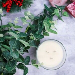 May include: A white candle with three wicks in a clear glass jar. The candle is surrounded by green eucalyptus leaves.