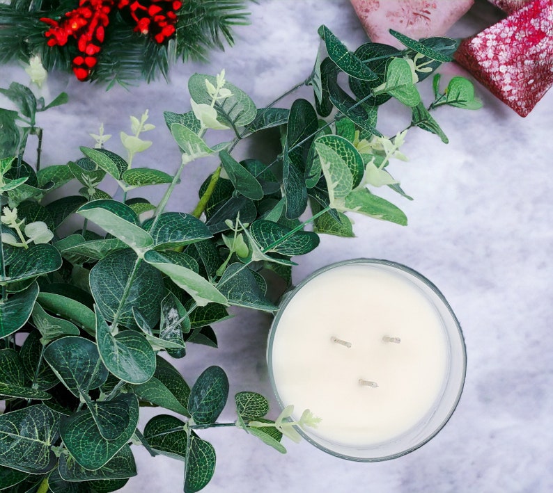 May include: A white candle with three wicks in a clear glass jar. The candle is surrounded by green eucalyptus leaves.