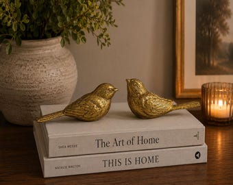 May include: Two gold-coloured bird figurines with textured feather details resting on a stack of two hardcover books. The top book is titled The Art of Home by Shea McGee, and the bottom book is titled This Is Home by Natalie Walton.