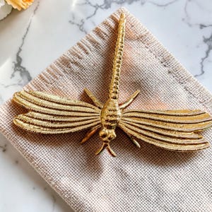 May include: A gold-colored dragonfly figurine with detailed wings and body. The dragonfly is resting on a beige napkin. The wings have horizontal lines, and the body has a textured design. The figurine is a decorative item.