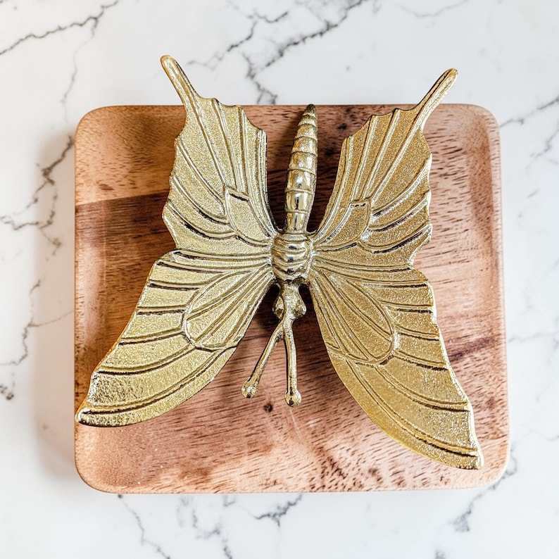 May include: A gold-colored butterfly-shaped decorative object with detailed wing patterns, resting on a square wooden tray. The butterfly has intricate detailing and is made of a shiny, metallic material. The tray has a natural wood grain finish.