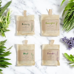 May include: Four beige burlap soap wash mitts with brown labels. The labels read "Rosemary," "Lemongrass," "Mint," and "Lavender." Each label has a botanical illustration and the net weight is 4 oz / 113g. Green and purple plants surround the mitts.