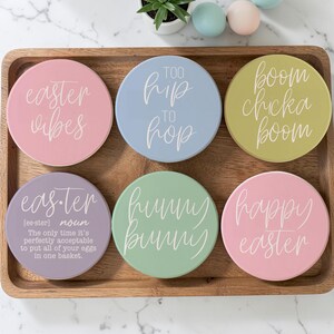 May include: A set of six pastel-colored ceramic coasters with Easter-themed phrases in white script. The phrases include "Easter Vibes," "Too Hip to Hop," and "Boom Chicka Boom." The coasters are arranged on a wooden tray.