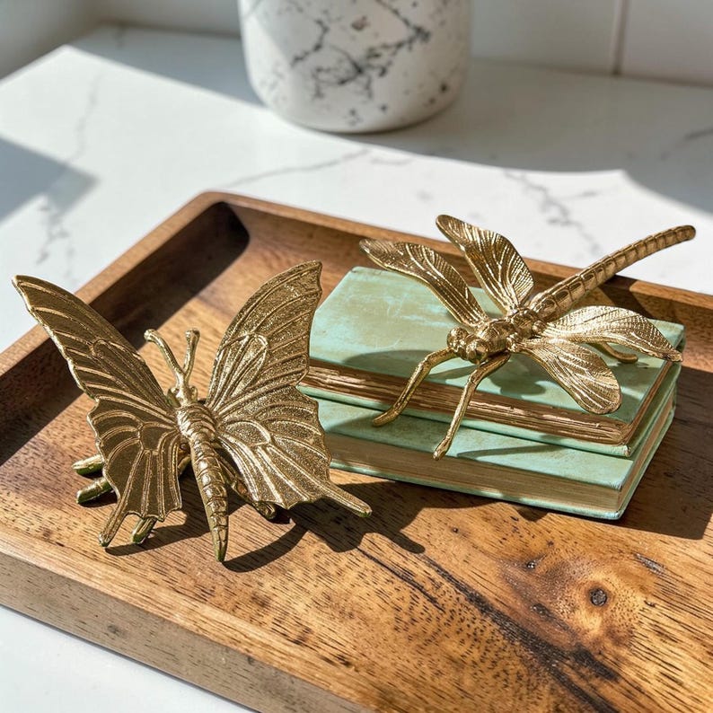 May include: A gold-colored butterfly and dragonfly figurine set. The butterfly is on the left, and the dragonfly rests on a stack of small, light-green books. The figurines are on a wooden tray.