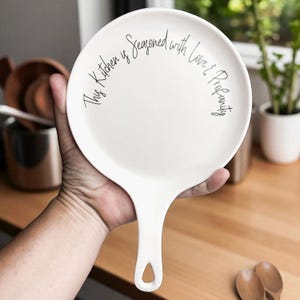 May include: White ceramic spoon rest in the shape of a frying pan. The spoon rest has a handle and a circular surface with the text "This Kitchen is Seasoned with Love & Prosperity" in black script.