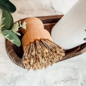 May include: A wooden dish brush with natural bristles, sitting in a wooden dish with a white bottle.