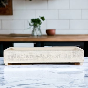 May include: A white wooden tray with the text "gather here with grateful hearts" on the front. The tray is sitting on a white marble surface.