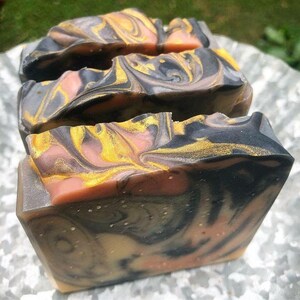 May include: Three bars of handmade soap with a swirl pattern of black, pink, and gold. The soap has a speckled appearance.