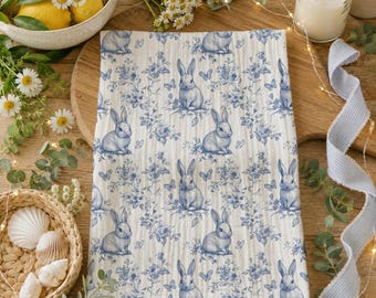 May include: A rectangular white kitchen towel featuring a repeating blue toile-style pattern of sitting rabbits, butterflies, and floral sprigs. The fabric has a textured, crinkled appearance, and the print is consistent across the entire surface.