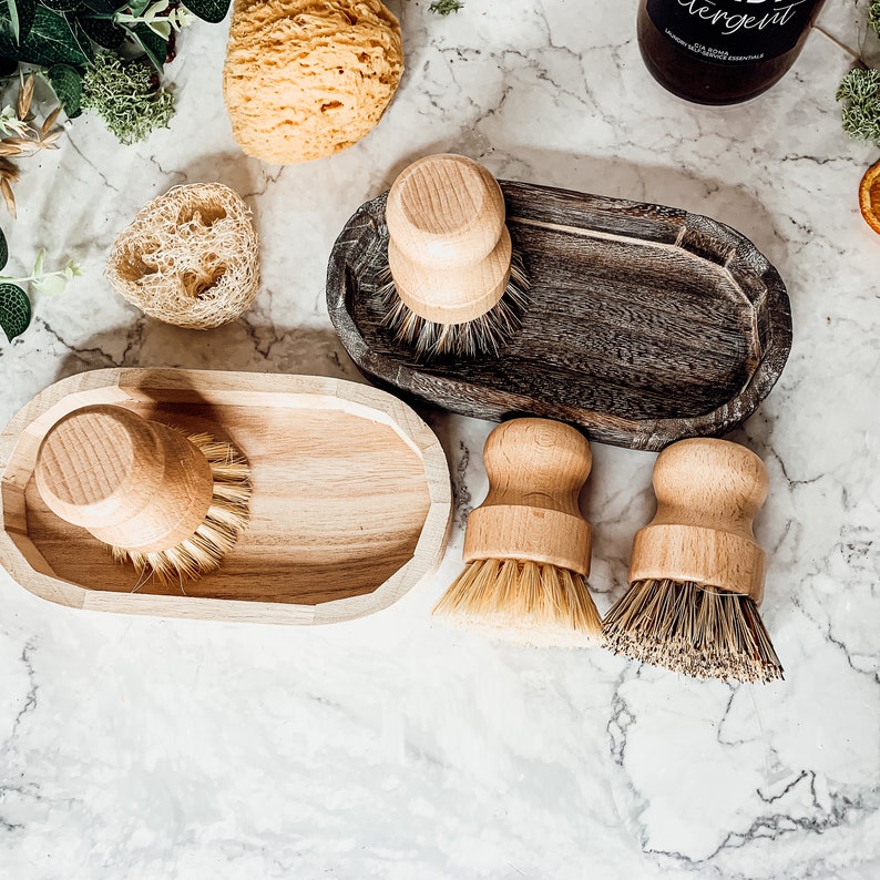 May include: Four wooden dish brushes with natural bristles in various sizes. The brushes are displayed in two wooden and one grey oval-shaped holders.