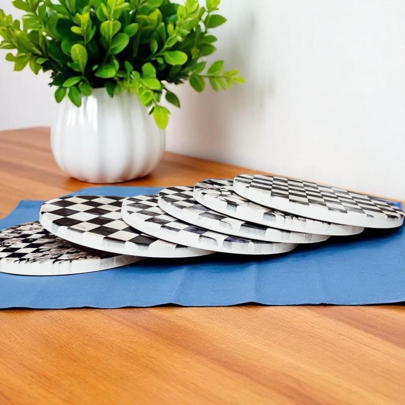 Op de afbeelding: Een set van zes ronde onderzetters met een zwart-wit dambordpatroon. De onderzetters liggen op een blauwe doek, met een witte vaas en een groene plant op de achtergrond. Een decoratief item.