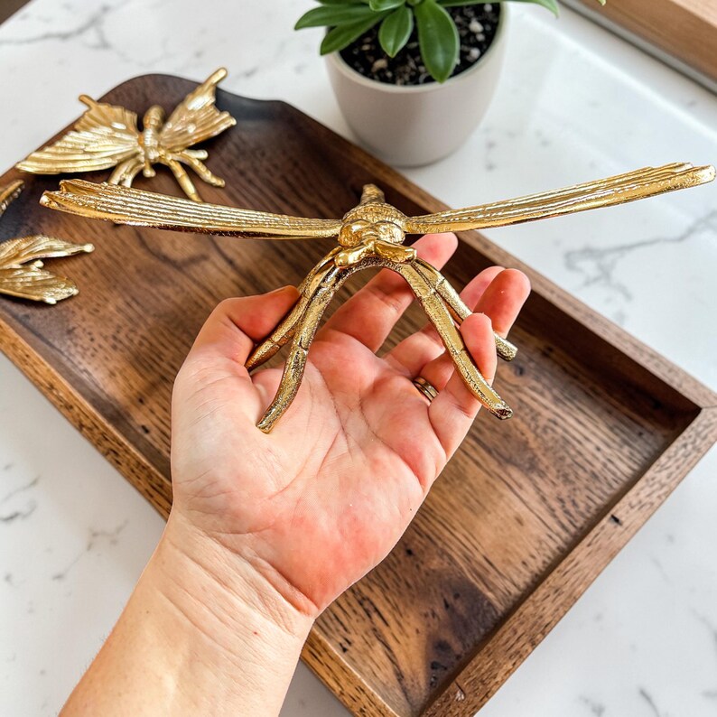 May include: A gold-colored dragonfly sculpture with outstretched wings is held in a hand. The sculpture is on a wooden tray, with a butterfly sculpture and a small potted plant in the background. The dragonfly is a decorative item.