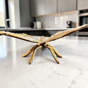 May include: A gold-colored metal butterfly sculpture with outstretched wings and detailed legs. The decorative piece is placed on a white countertop, showcasing its intricate design and metallic finish. The sculpture is a decorative accent.