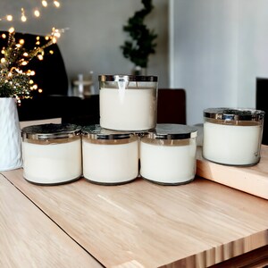 May include: Five white candles in glass jars with silver lids. The candles are arranged in a row on a wooden surface.