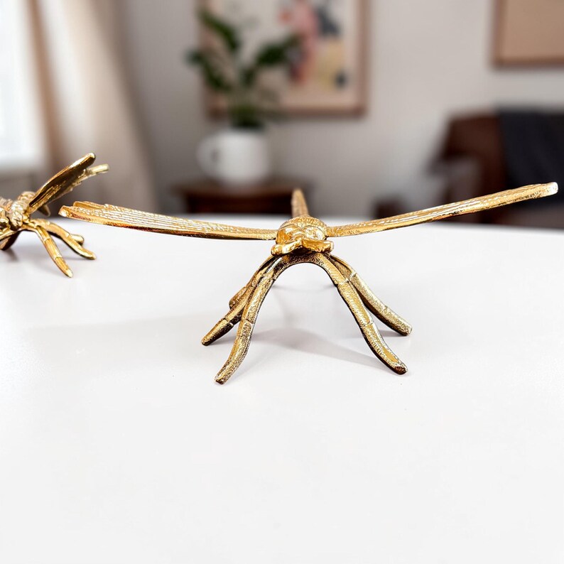 May include: Two gold-colored dragonfly sculptures. The dragonflies have outstretched wings and are positioned on a white surface. The sculptures have detailed legs and bodies, and are made of a metallic material.