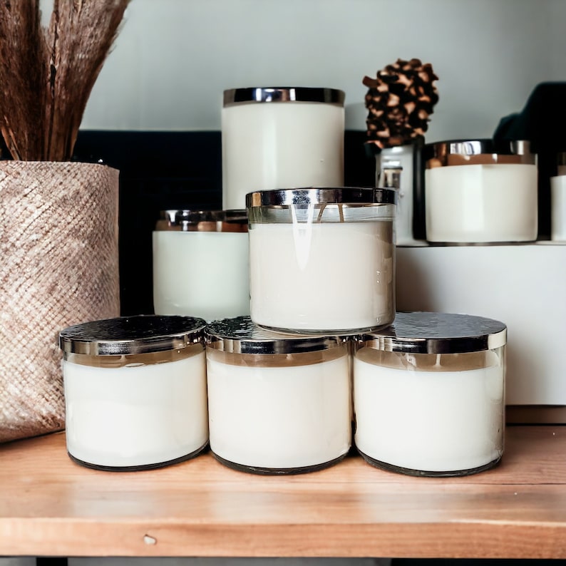 May include: A collection of six glass candle jars with silver lids. The jars are filled with white candles and are arranged in a pyramid shape on a wooden surface.