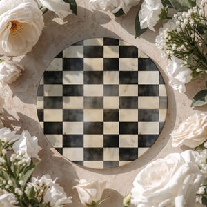 Op de afbeelding: Een ronde decoratieve schaal met een zwart-wit geblokt patroon. De schaal is omgeven door witte rozen en andere witte bloemen, waardoor een bloemenrand ontstaat. De schaal wordt waarschijnlijk gebruikt om te serveren of te presenteren.