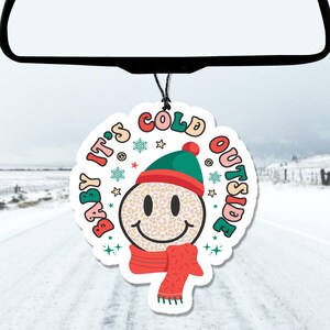 May include: A white, circular car air freshener with a smiling face wearing a green and red hat and a red scarf. The text "BABY IT'S COLD OUTSIDE" is written around the smiley face in various colors and fonts. The background shows a snowy road.