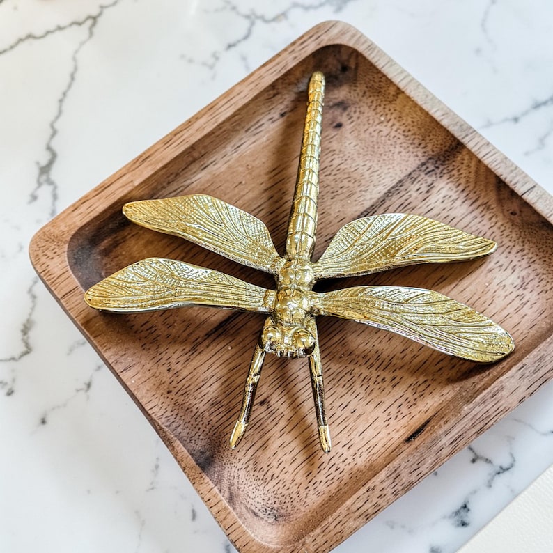 May include: A gold-colored dragonfly figurine with detailed wings rests in a square, wooden tray. The dragonfly has a long, segmented body and delicate legs. The tray has rounded corners and a natural wood grain.