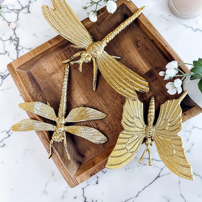May include: Three gold-colored decorative insect figurines: two dragonflies and one butterfly. The insects are placed on a square wooden tray. The dragonflies have detailed wings and bodies. The butterfly has detailed wings and antennae.