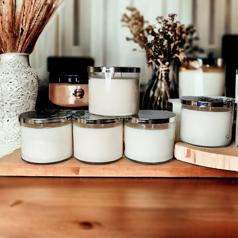 May include: Five white candles with silver lids are arranged on a wooden surface. The candles are in various sizes and are all lit.