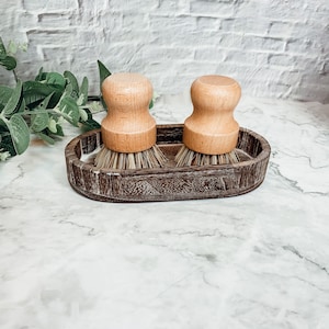 May include: Two wooden handled scrub brushes with natural bristles are displayed in a dark brown wooden dish. The dish has a slightly distressed finish.