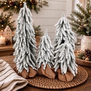 May include: Three artificial Christmas trees of varying heights, each featuring dense green needles heavily dusted with white flocking to resemble snow. The base of each tree is wrapped in a natural brown burlap fabric tied with twine.