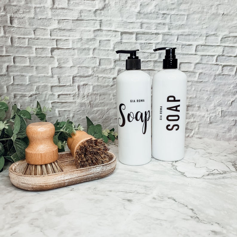 May include: Two white soap pump bottles with black pumps and black text that reads "Soap" and "Gia Roma" on a white countertop.  Two wooden scrub brushes are in a wooden tray.