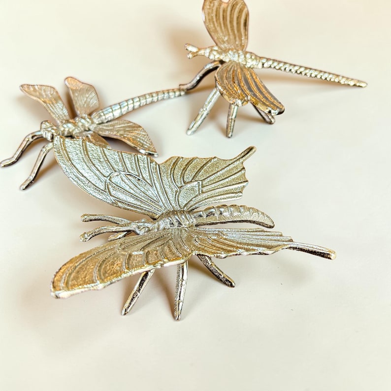 May include: Three silver-colored metal insect figurines: two dragonflies and one butterfly. The dragonfly figurines have long, slender bodies and delicate wings. The butterfly has large, detailed wings. All have intricate detailing and are displayed on a light background.