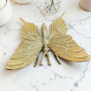 May include: A gold-colored butterfly-shaped decorative object. The butterfly has detailed wing patterns and a textured body. The wings are spread wide, and the object is resting on a white marble surface.